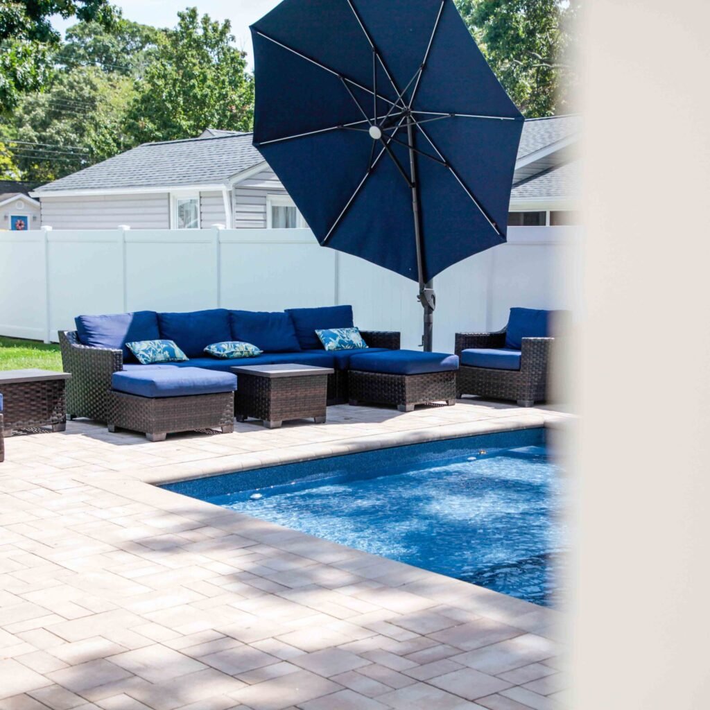 Rectangular in-ground pool with dark blue water, viewed from the paver patio. A wicker sectional sofa and chair set with navy blue cushions and a large navy market umbrella are arranged on the tan paver deck adjacent to the pool. A white vinyl privacy fenc
