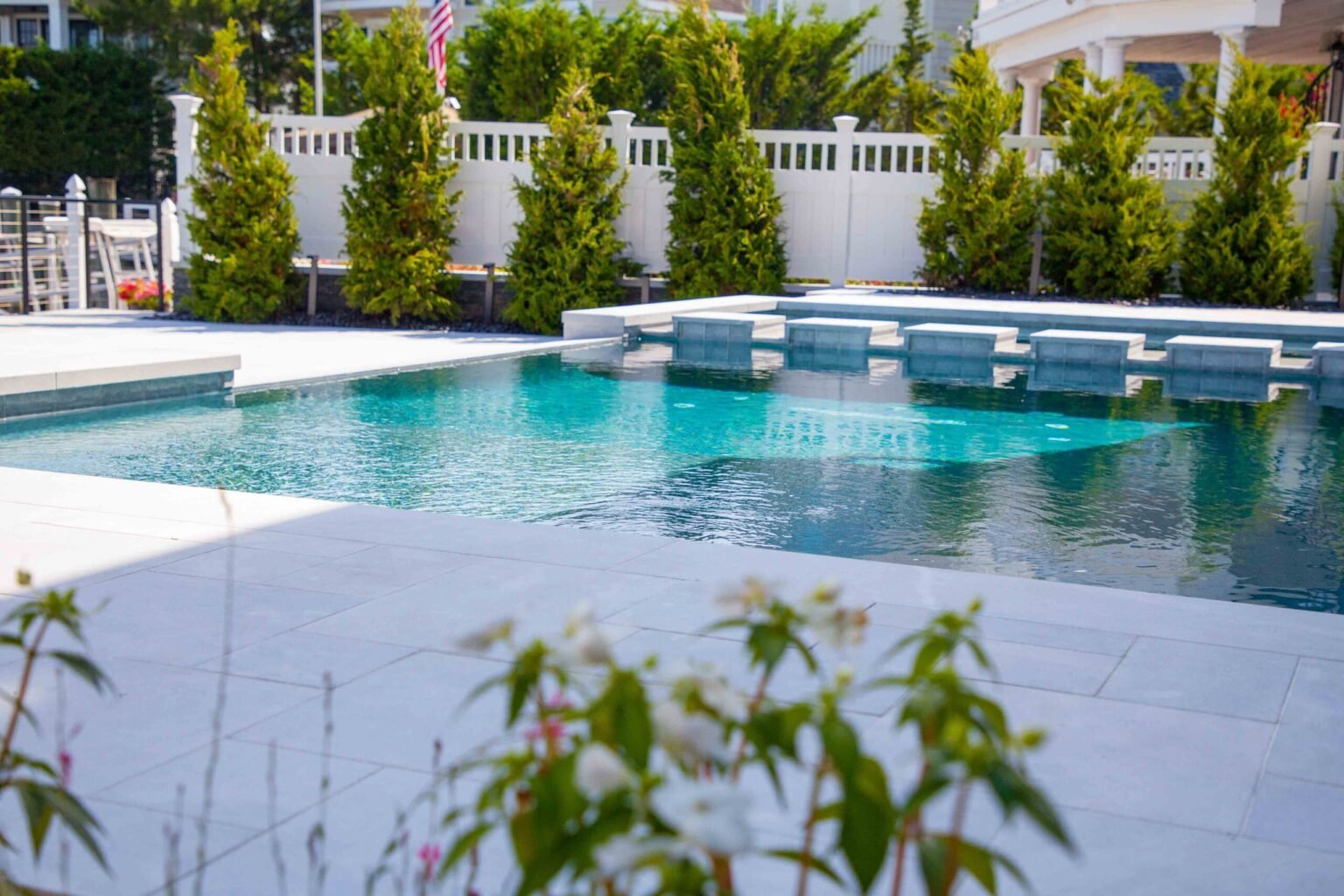 Rectangular in-ground pool with wide tanning ledge entry steps and light gray tile, surrounded by large-format white paver decking, a white vinyl privacy fence, and a row of mature evergreen arborvitae plantings.