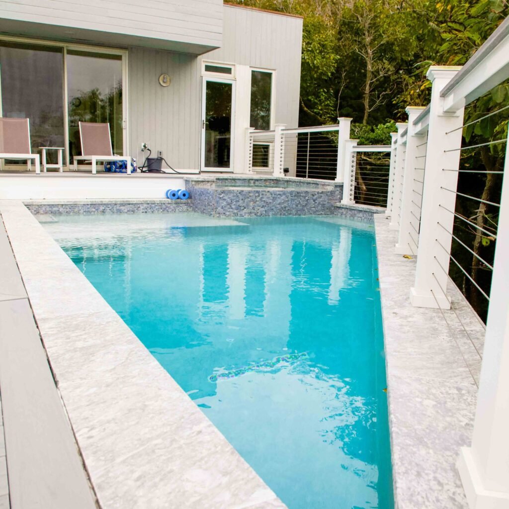 Rectangular in-ground pool with a gray mosaic tile waterline border and travertine coping and deck, adjacent to a modern white-sided home with sliding glass doors. A white cable-rail fence lines one side of the pool, and lounge chairs are positioned on the