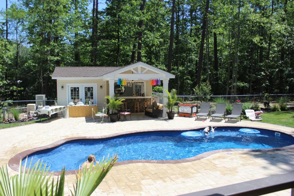 Freeform in-ground pool with deep blue water and a tan paver surround, set in a wooded backyard. A white pool house with a covered outdoor bar and lounge area is visible behind the pool, with lounge chairs and tropical potted plants arranged on the patio.