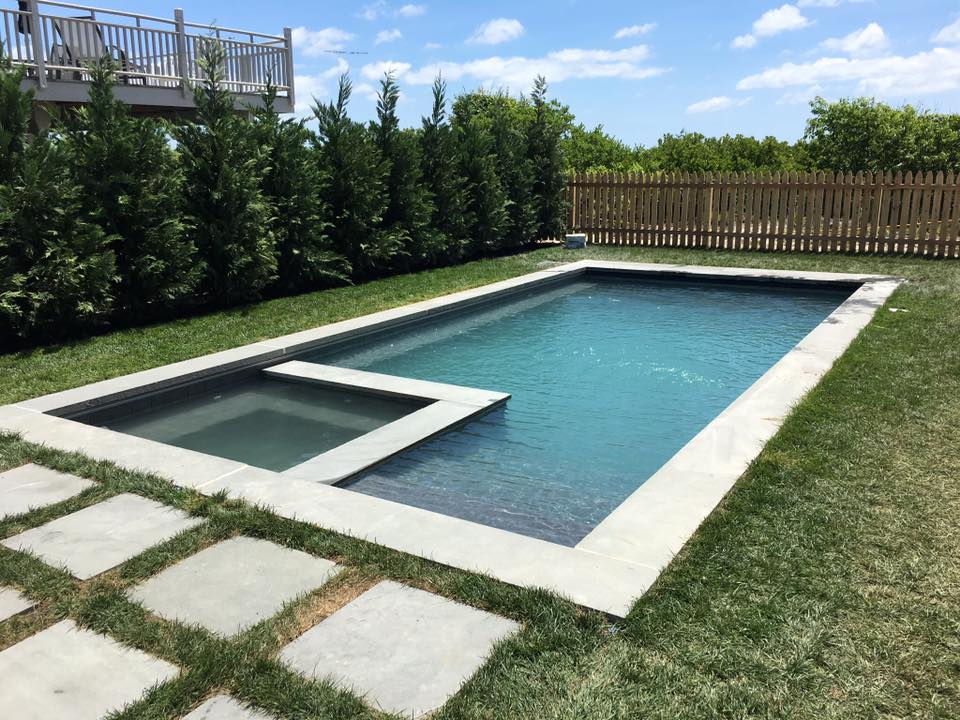 An aerial view of a freeform in-ground pool with a natural rock waterfall feature at the far end. The pool is surrounded by a stamped concrete patio in a tan and brown tone. A black aluminum fence with an arched double gate encloses the pool area. A small 