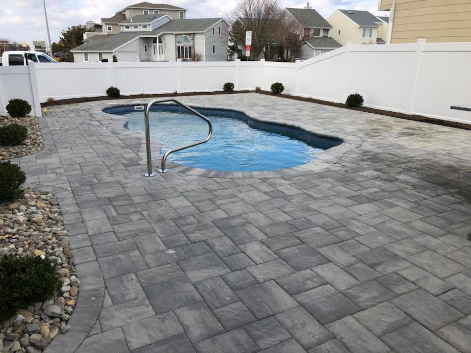 Freeform in-ground pool with bright blue water and a stainless steel handrail, set within a large gray paver deck. A white vinyl privacy fence borders the yard, with neighboring homes visible beyond.