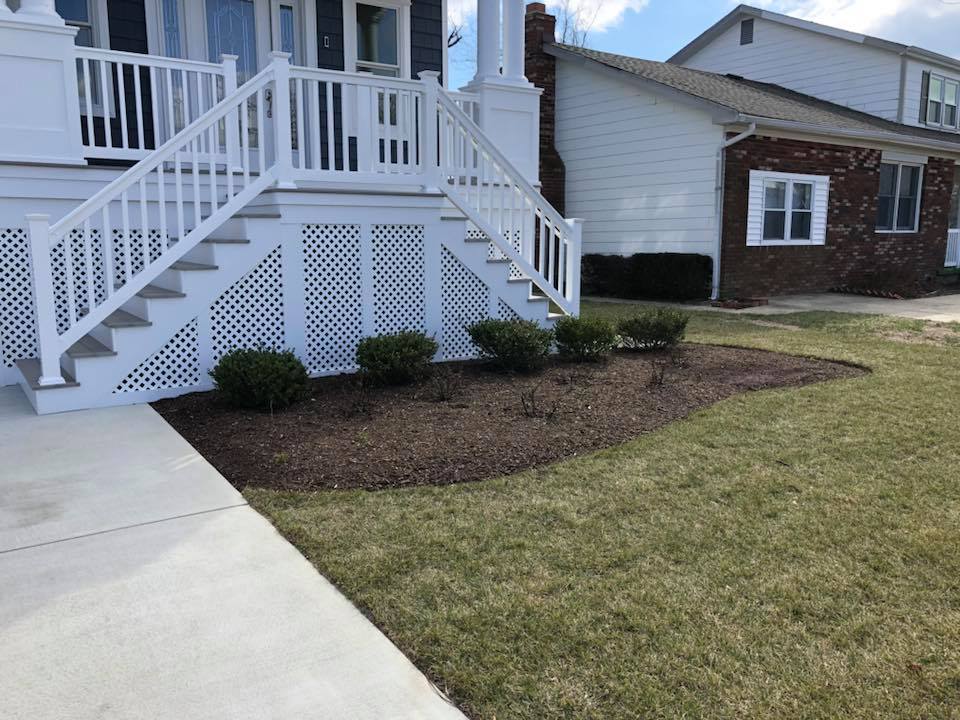 White vinyl front porch staircase with lattice skirting, flanked by a freshly mulched landscape bed planted with small shrubs along a concrete walkway.