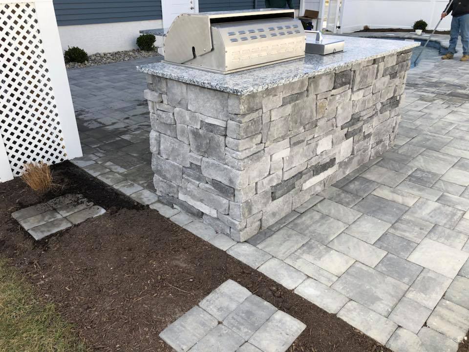 Construction photo of a stacked-stone outdoor kitchen island with a granite countertop and built-in grill on a newly installed gray concrete paver patio. Landscaping around the patio is incomplete, and a worker is visible in the background near the pool ar