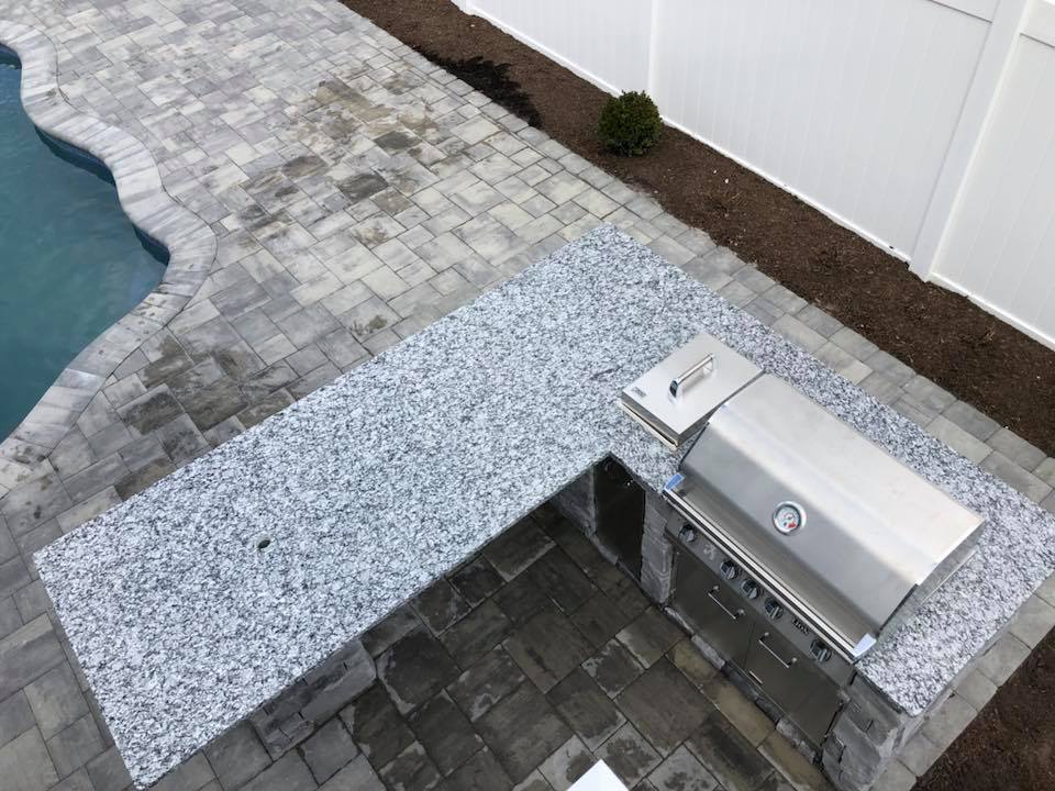 Overhead view of an L-shaped outdoor kitchen built on a gray paver patio, featuring a granite countertop and a stainless steel built-in grill. The edge of a freeform pool with paver coping is visible in the upper left corner of the frame.