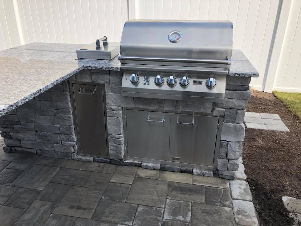 L-shaped outdoor kitchen built with gray stone veneer and a granite countertop, featuring a stainless steel built-in grill with multiple burner knobs and stainless steel access doors below. The unit sits on a gray paver patio with a white vinyl fence in th