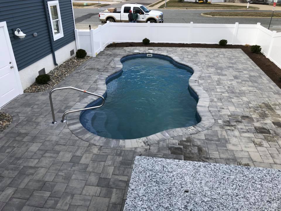 Overhead view of a freeform kidney-shaped in-ground pool with a dark blue liner and gray paver coping, surrounded by a gray paver deck. A stainless steel handrail marks the entry point, and a white vinyl fence encloses the backyard next to a blue-sided hou