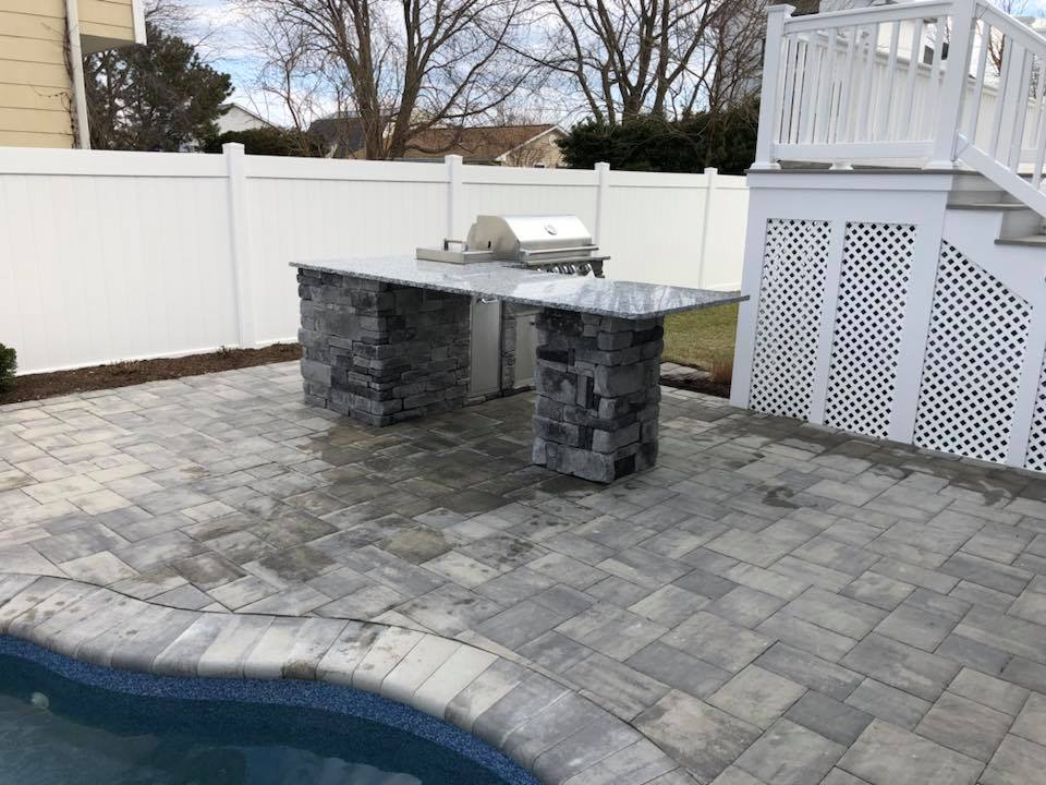 Stacked-stone outdoor kitchen island with a granite countertop and built-in stainless steel grill sits on a gray concrete paver patio adjacent to the curved edge of an in-ground pool. A white vinyl privacy fence and white lattice deck skirting frame the ba
