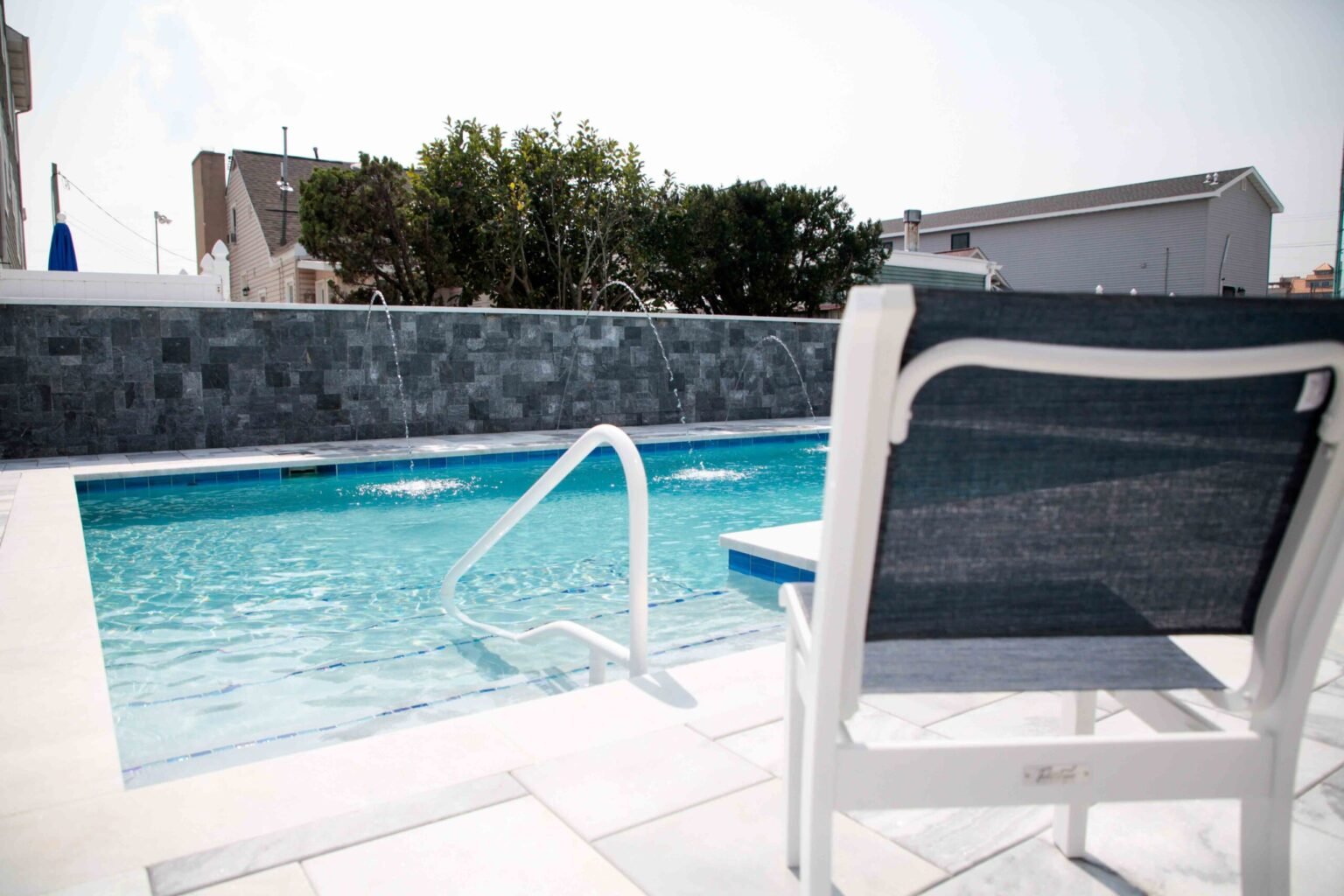 Rectangular in-ground pool with light blue water, photographed from a white paver deck at water level. Three sheer descent water jets arch from a dark gray stone block wall into the pool. A white lounge chair and stainless steel entry rail are visible in t