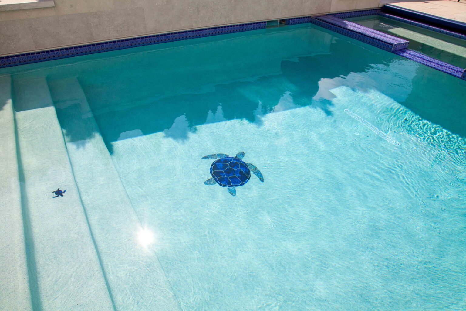 Close-up of the interior floor of a pool with crystal-clear light blue water showing two blue ceramic mosaic sea turtle inlays on the white pool floor. A blue tile waterline border and wide entry step ledge are visible along the edges. A separate attached 