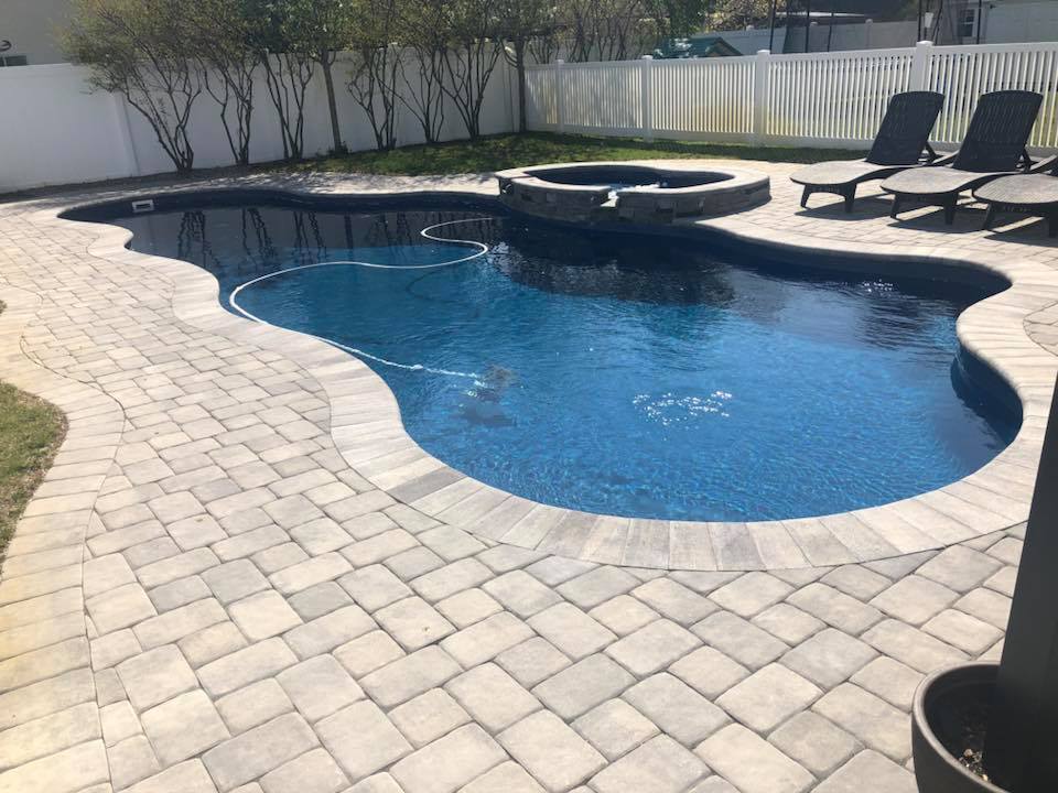 Freeform in-ground pool with a dark blue liner and an attached raised round spa with stone coping, set on a large gray paver deck. Dark lounge chairs sit on the deck, and a white vinyl picket fence with bare trees lines the perimeter of the yard.