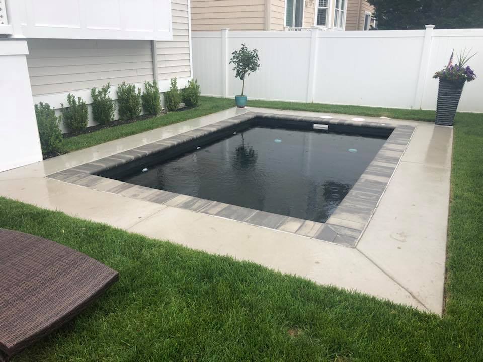 Small square in-ground pool with a dark charcoal liner and stone tile coping, surrounded by a concrete deck and green lawn. A white vinyl privacy fence encloses the backyard, and trimmed shrubs line the adjacent house foundation.