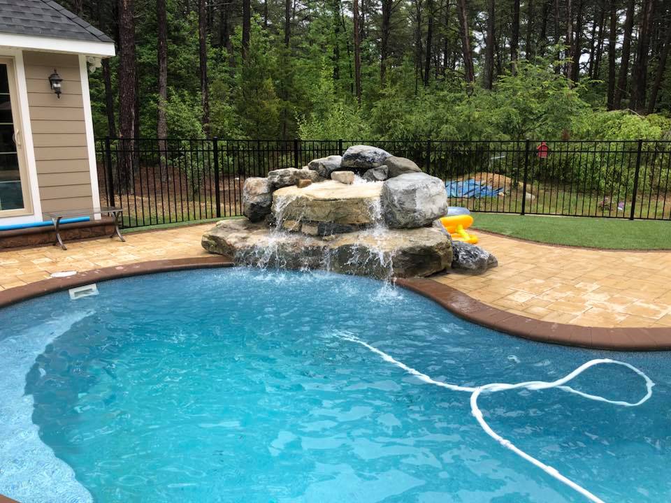 Freeform in-ground pool with a large natural boulder waterfall cascading into the water. Tan paver coping and deck surround the pool, a black aluminum fence lines the perimeter, and a wooded tree line fills the background.