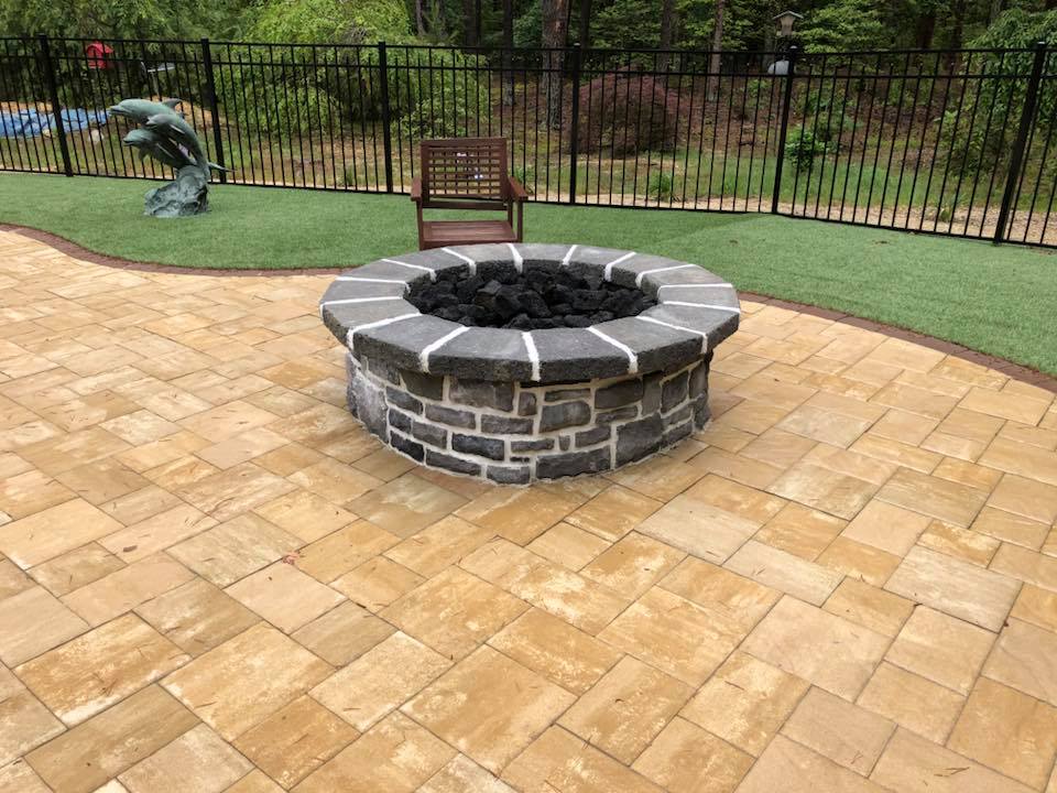 Circular stone fire pit with lava rock fill, built with gray stacked stone and a granite cap, centered on a large tan paver patio. A green lawn with artificial turf, a dolphin sculpture, and a black aluminum fence surround the space.