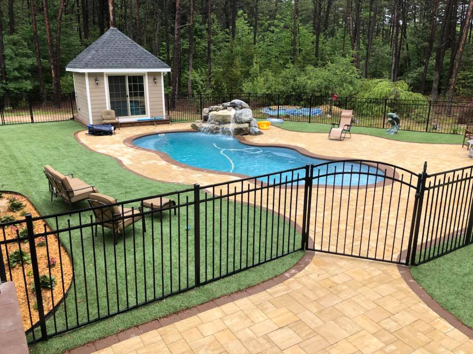 A rectangular in-ground pool with an attached square spa in the near left corner, both finished with dark gray plaster and gray bluestone coping. The pool is set in a grass lawn enclosed by a wood picket fence and a row of tall evergreen arborvitae. A whit