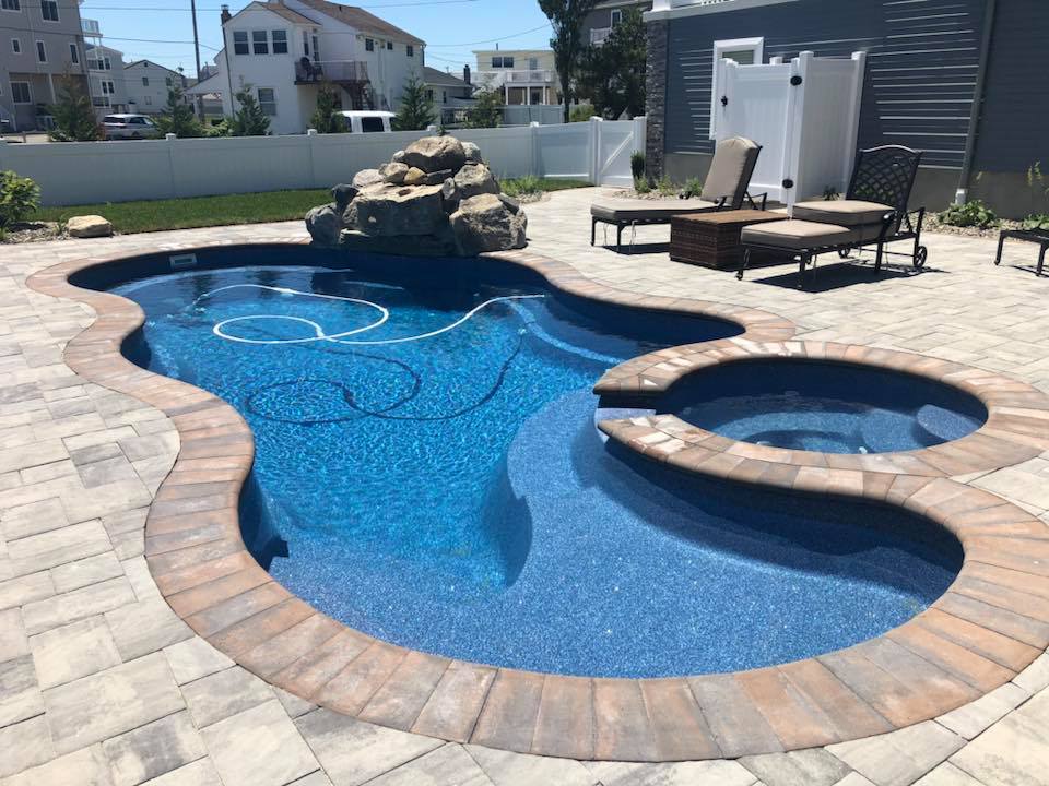Freeform in-ground pool with a dark blue liner and an attached round spa, connected by a warm-toned paver coping and deck. A natural rock waterfall feature rises at the far end of the pool, and lounge chairs are positioned nearby within a white vinyl fence