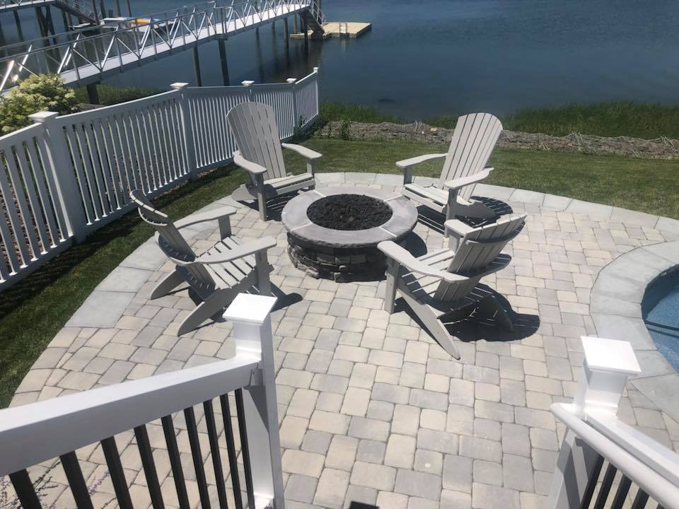 Tan brick paver patio featuring a round stacked-stone fire pit surrounded by four gray Adirondack chairs. White vinyl railing borders the patio; a dock and open waterway are visible in the background. The partial edge of a pool with blue water is visible a