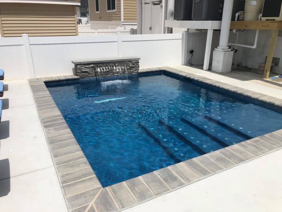 Rectangular in-ground pool with deep blue water and a stacked-stone spillover water feature along the back wall. The pool has gray paver coping, a concrete deck surround, visible entry steps, and is enclosed by a white vinyl privacy fence.