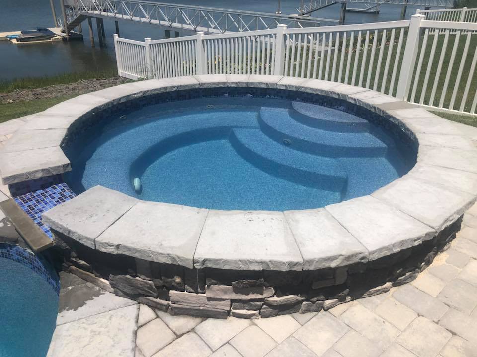 Circular raised in-ground spa with a stacked-stone exterior wall, gray paver coping, and a blue mosaic tile accent strip at the waterline. The spa sits on a paver patio adjacent to a waterway with a dock visible behind a white vinyl railing fence.