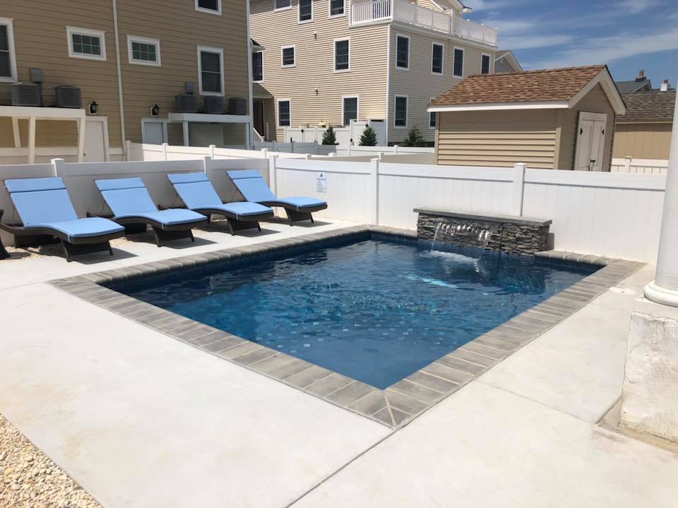 Rectangular in-ground pool with gray large-format concrete coping and a gray stacked-stone waterfall spillway on the far end. A concrete deck surrounds the pool with four dark-framed lounge chairs with blue cushions along one side. The backyard is enclosed