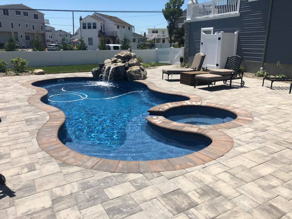 Freeform in-ground pool with a dark blue liner and an attached circular spa, surrounded by large-format tan and gray pavers. A natural boulder waterfall sits at the far end of the pool, and lounge chairs are arranged on the paver deck within a white vinyl 