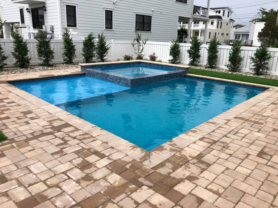 Rectangular in-ground pool with a raised square spa built into the pool interior and tiled in iridescent blue-gray mosaic tile. Tan brick paver coping and a paver deck surround the pool. A white vinyl privacy fence lines the yard perimeter, with a row of a