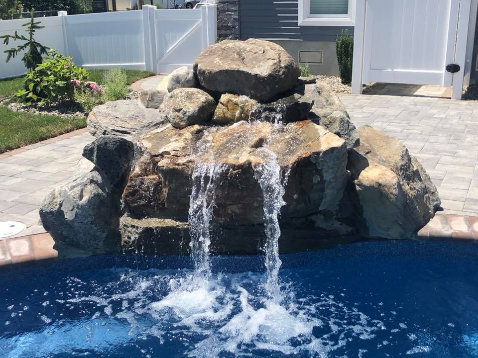 Large natural boulders stacked to form a waterfall feature, with water cascading down the rock face into a dark-blue in-ground pool. A tan paver patio deck is visible at the base of the rocks, with a white vinyl fence and gate in the background.