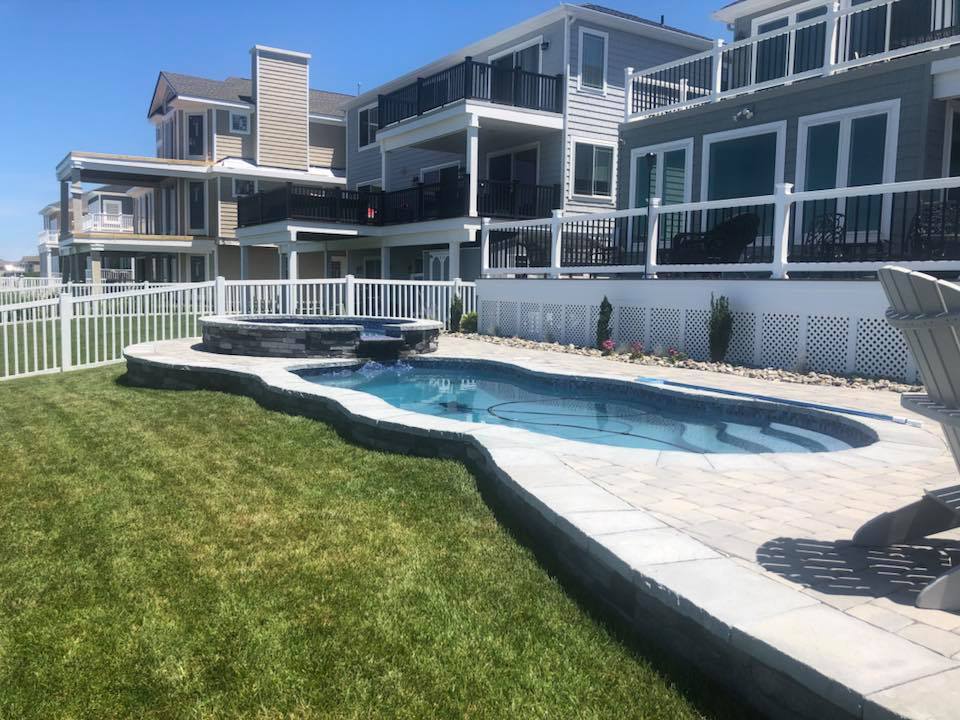 Freeform in-ground pool with an attached raised circular spa, finished with light gray concrete coping. A manicured green lawn borders the pool on one side, a white vinyl fence encloses the yard, and multi-story coastal homes are visible in the background.