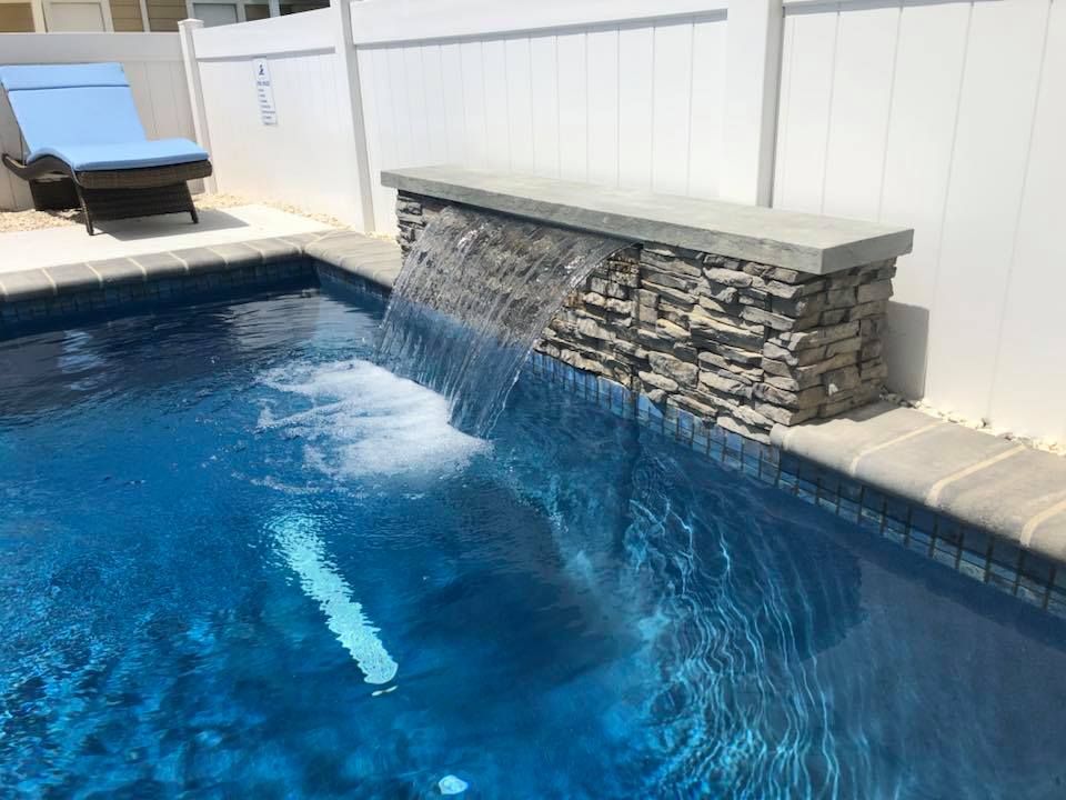 Close-up of an in-ground pool featuring a stacked-stone waterfall wall with a bluestone cap, cascading water into the pool. Blue mosaic tile lines the waterline, concrete coping borders the edge, and a white vinyl privacy fence encloses the backyard.