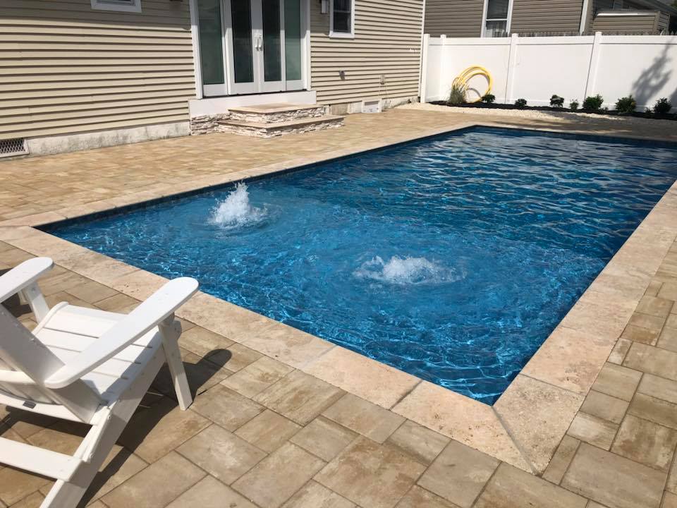 Rectangular in-ground pool with a dark blue liner and two active in-pool bubbler fountains, surrounded by a tan stamped paver deck. A white Adirondack chair sits in the foreground, and a raised stone step structure is visible near the house entry.