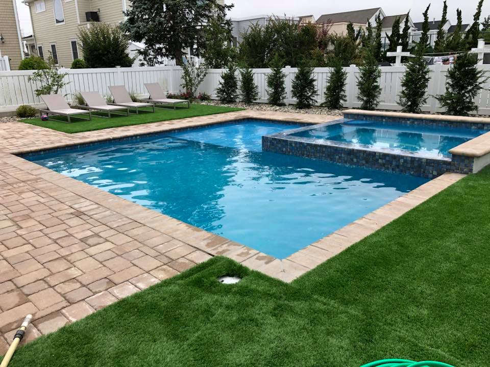 Rectangular in-ground pool with an elevated square corner spa faced in iridescent blue-gray mosaic tile. Tan brick paver coping and a paver deck surround the pool, with artificial turf filling the yard on two sides. Two lounge chairs sit on the turf, and a