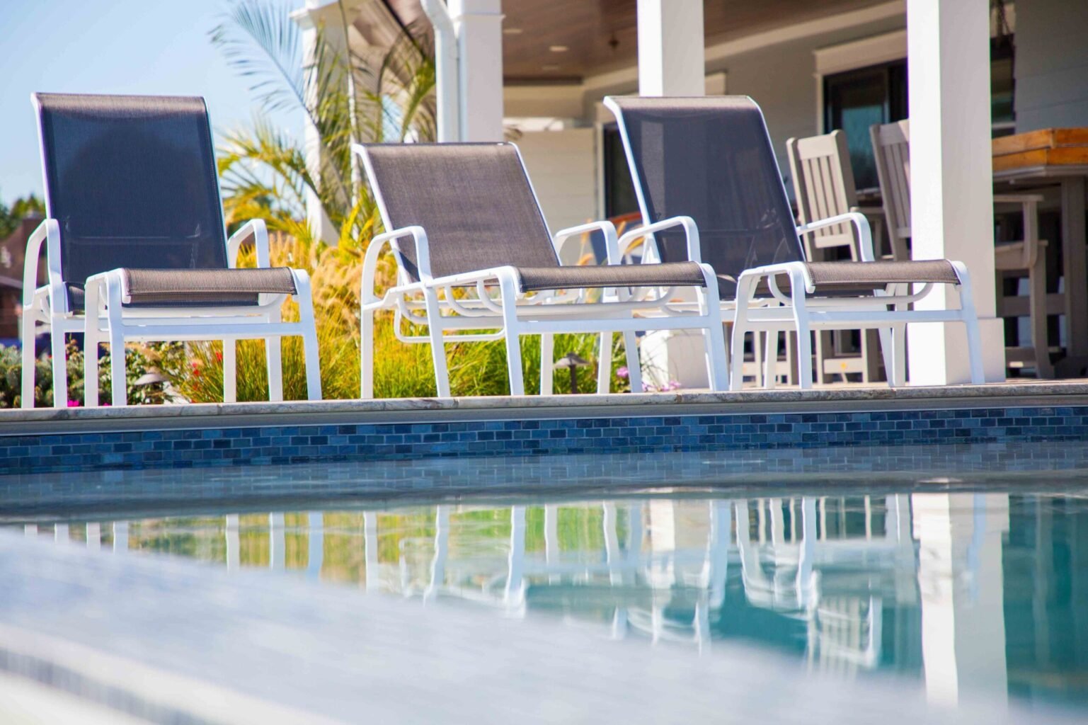 Ground-level view along the edge of a completed in-ground pool showing a row of four white-frame mesh sling lounge chairs positioned on a light gray concrete pool deck. Dark blue mosaic waterline tile runs along the pool wall at deck level. A white-columne