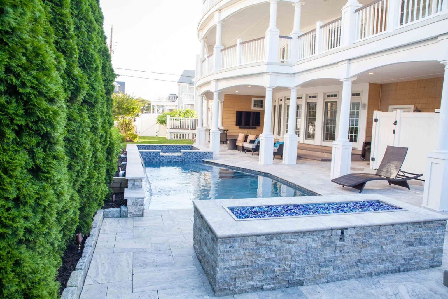 A narrow rectangular lap pool with blue glass mosaic waterline tile runs alongside a large multi-story coastal home featuring white columns and a wraparound upper balcony. A stone-clad rectangular fire pit filled with blue glass fire media sits in the fore
