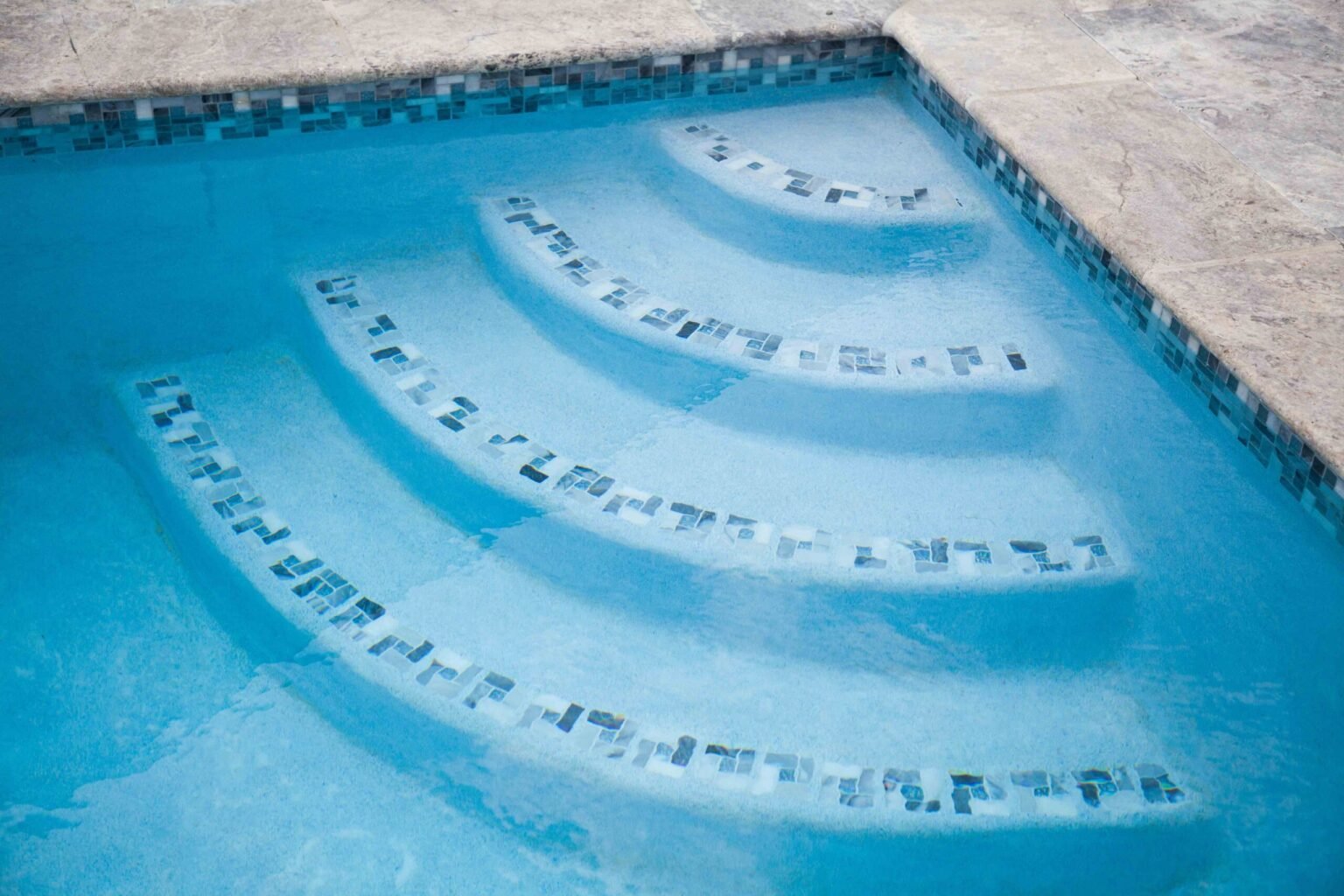 Close-up of a completed in-ground pool interior showing curved, semicircular entry steps submerged underwater. Each step is edged with a row of small iridescent mosaic tiles in blue and silver tones. The surrounding coping is natural travertine, and the po