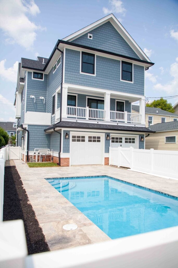 Rectangular in-ground pool with clear blue water set against the rear of a three-story blue-gray shingle-style home. The surrounding deck is finished in travertine pavers. A white vinyl privacy fence runs along the property sides, and a two-car garage with