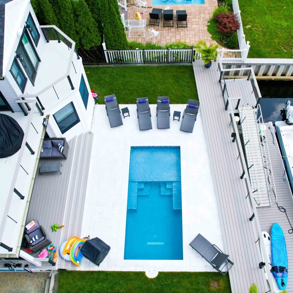 Overhead aerial view of a rectangular in-ground pool centered on a composite deck between a white two-story coastal home and a boat dock, with gray lounge chairs arranged poolside, outdoor living furniture, and a manicured lawn visible beyond a white fence