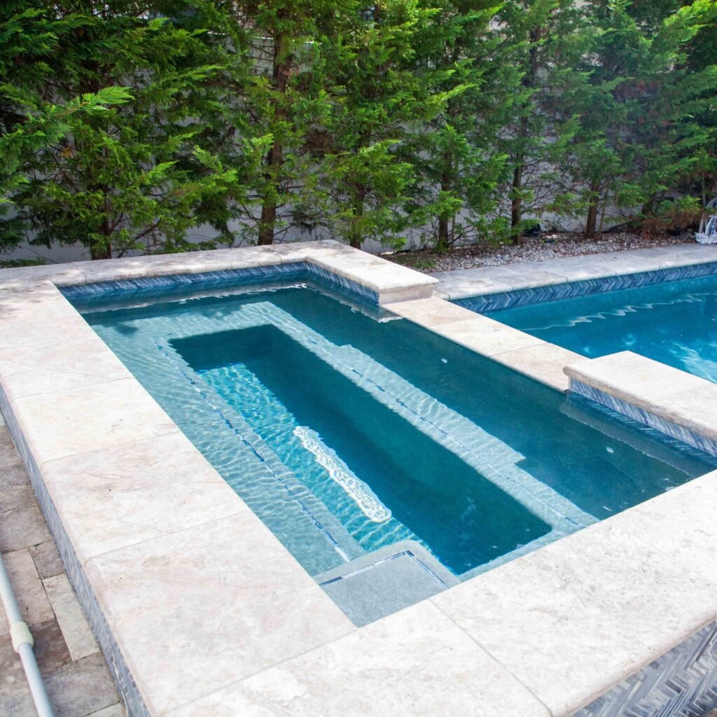 Overhead-angle view of a rectangular in-ground pool with an attached rectangular spa, both finished in dark blue plaster. The deck and coping are cream-colored travertine tile, and the backyard is enclosed by tall arborvitae trees and a stone retaining wal