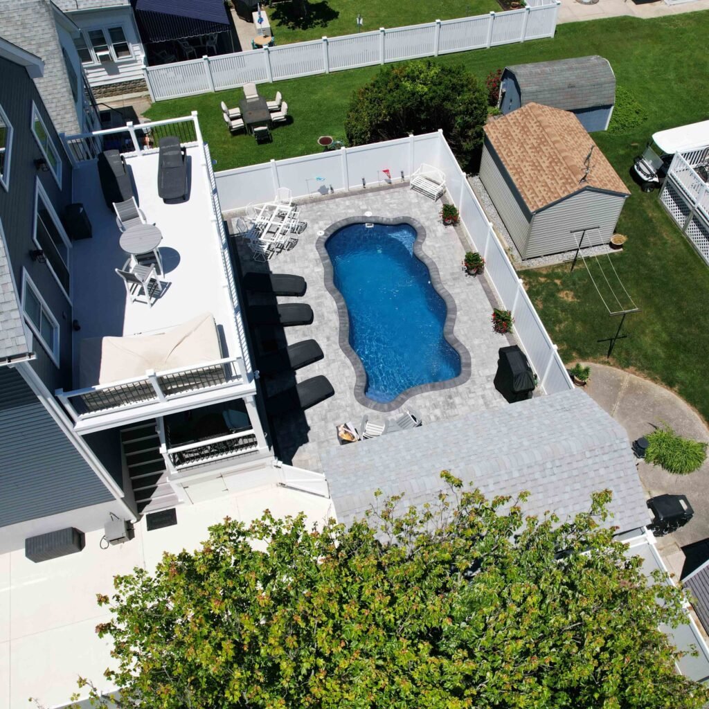 Overhead aerial view of a freeform in-ground pool with a scalloped shape, surrounded by a large gray paver patio and white vinyl privacy fence. The backyard includes lounge chairs, a gas grill, and a multi-level deck attached to the home.