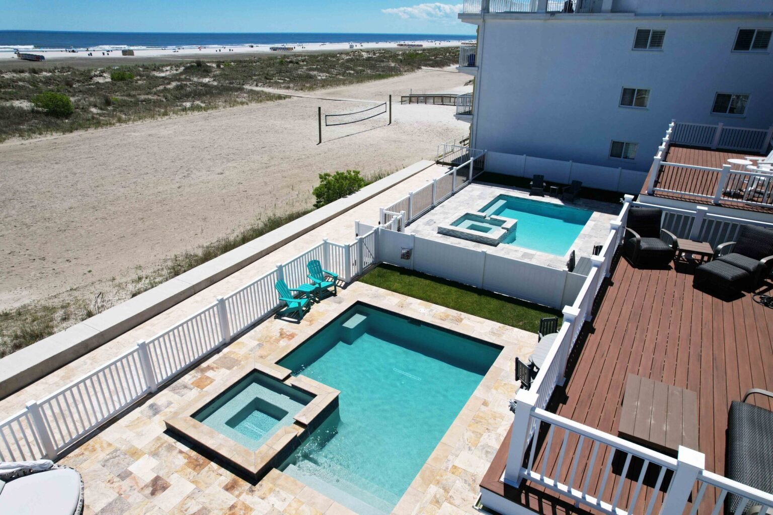 An aerial view of two rectangular in-ground pools with attached spas situated adjacent to a multi-story building, separated by a white vinyl fence. The pool decks are paved in travertine, with a small grass area and two blue Adirondack chairs visible. A sa