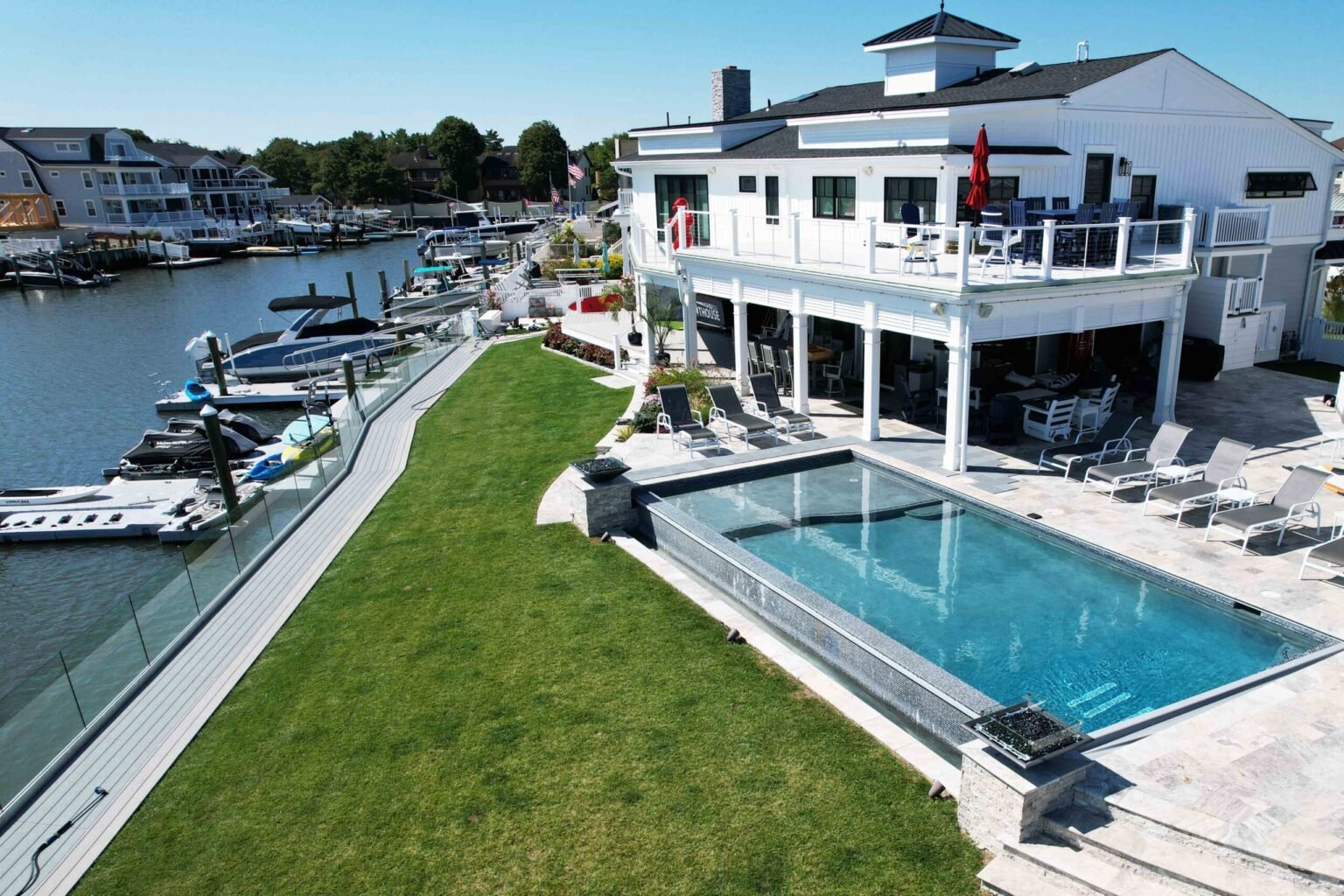 A large rectangular in-ground pool with a perimeter-overflow or infinity edge sits on a travertine paver deck lined with lounge chairs, directly adjacent to a marina waterfront with docks and boats. A glass panel railing separates the lawn from the water. 