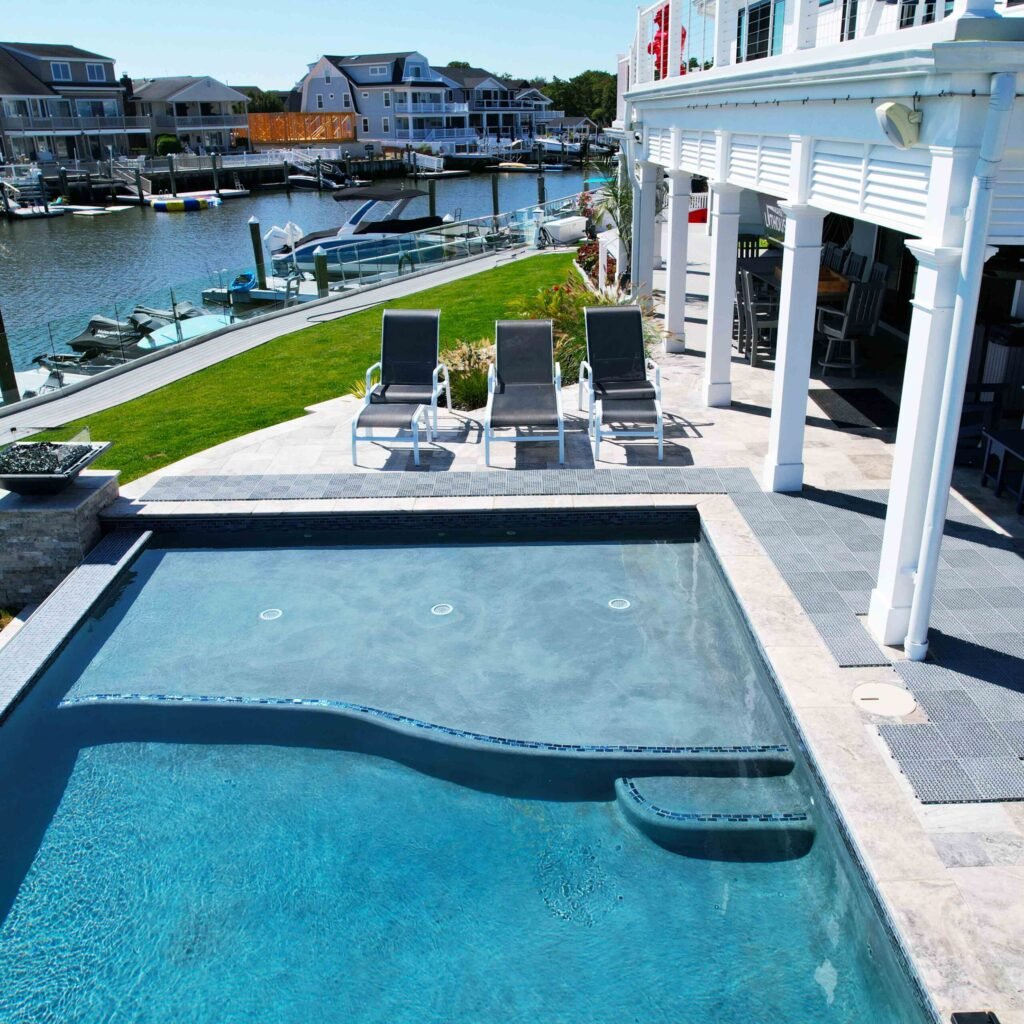 Elevated ground-level view of a rectangular in-ground pool and attached raised spa on a travertine paver deck, situated directly behind a waterfront home overlooking a marina with docks and boats. Three sling lounge chairs are positioned on the deck, and a