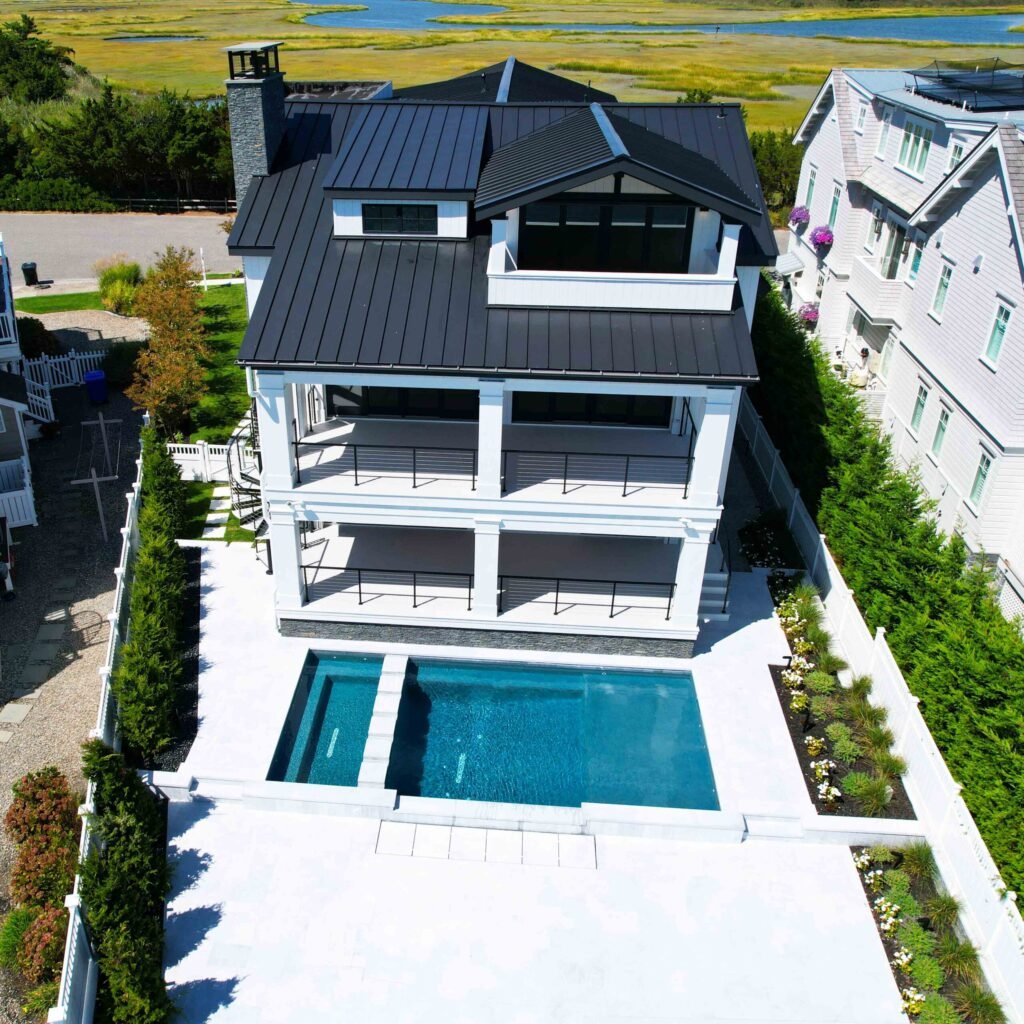 Aerial drone shot of a rectangular in-ground pool with an attached wading ledge set into a large white concrete deck behind a three-story coastal home with a black metal standing-seam roof. The property backs up to open salt marsh, with mature arborvitae h