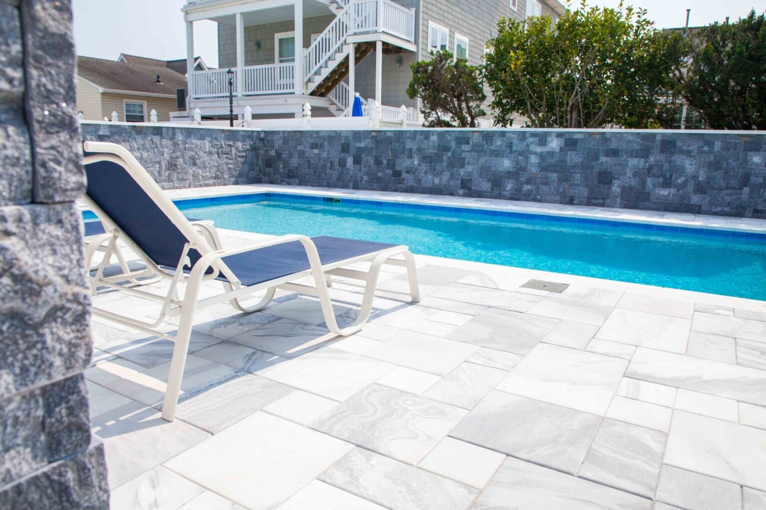 Rectangular in-ground pool bordered by a full-height gray stone block retaining wall and a large-format white marble-look porcelain paver deck. A blue-and-white lounge chair sits poolside, with a coastal residential neighborhood visible in the background.