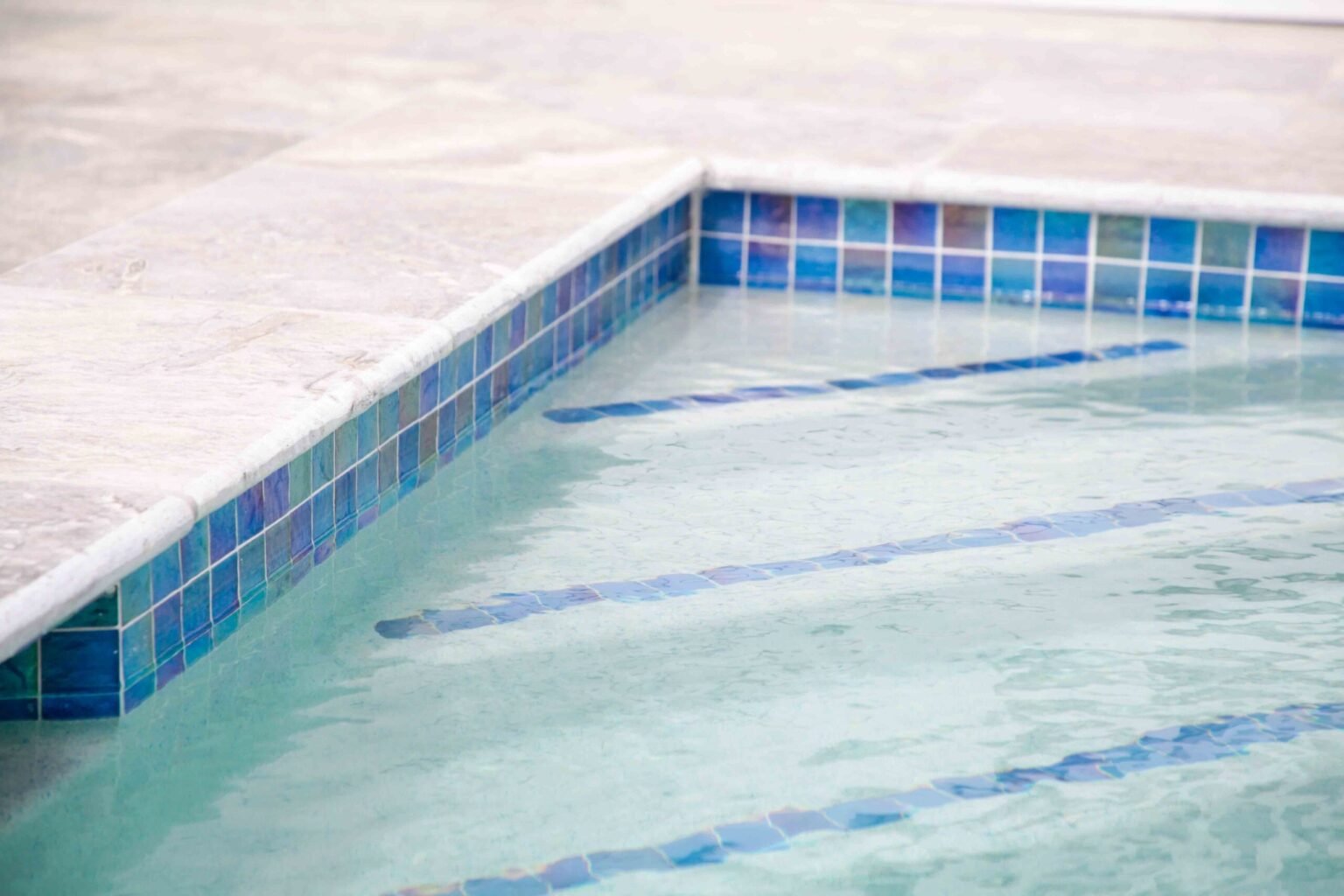 Close-up of a pool waterline showing iridescent blue glass mosaic tile with white grout, bordered by a white marble coping. A matching blue tile step strip is visible beneath the water surface.