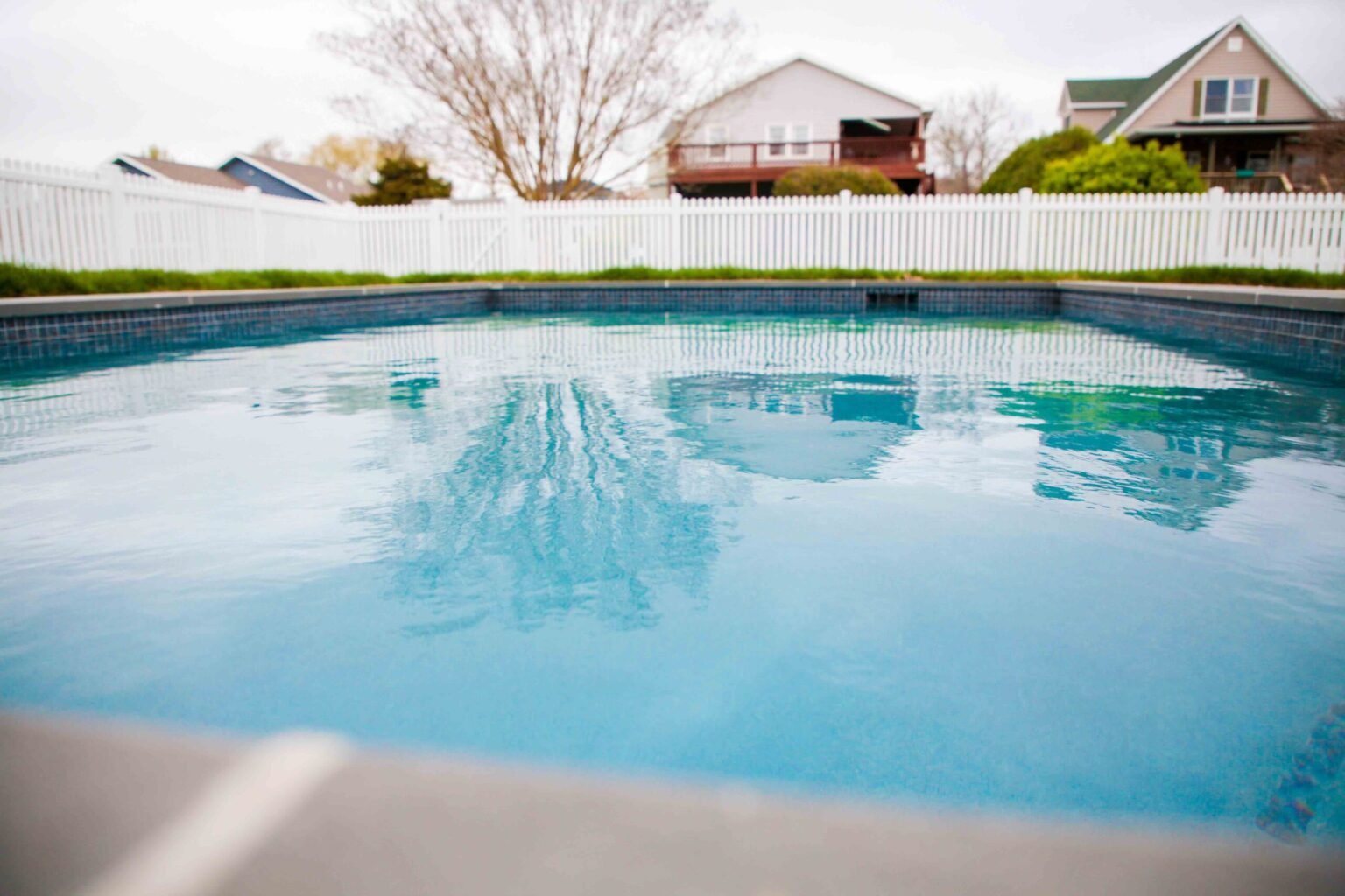 Rectangular in-ground pool filled with blue water, viewed from water level looking toward a white picket vinyl fence. Dark blue waterline tile lines the perimeter, and neighboring homes with a wood deck are visible beyond the fence in an overcast setting.