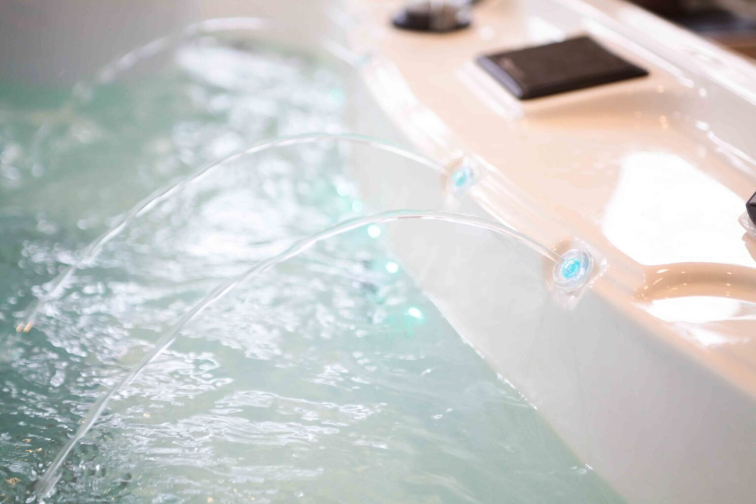 Close-up detail of multiple arc-shaped laminar water jets emerging from a white fiberglass shell and arching into light-blue water below. A blue LED light is visible beneath one jet nozzle; the image is tightly framed with a shallow depth of field.