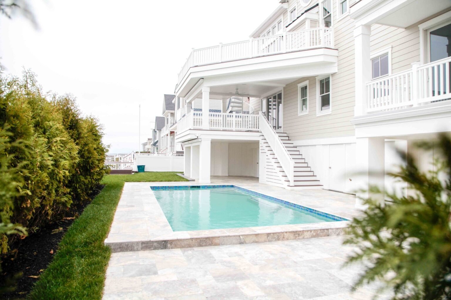 A completed rectangular in-ground pool set on a travertine paver deck adjacent to a large multi-story coastal-style home with white siding and wraparound balconies. The pool has a light blue interior finish and is surrounded by a manicured lawn with arborv