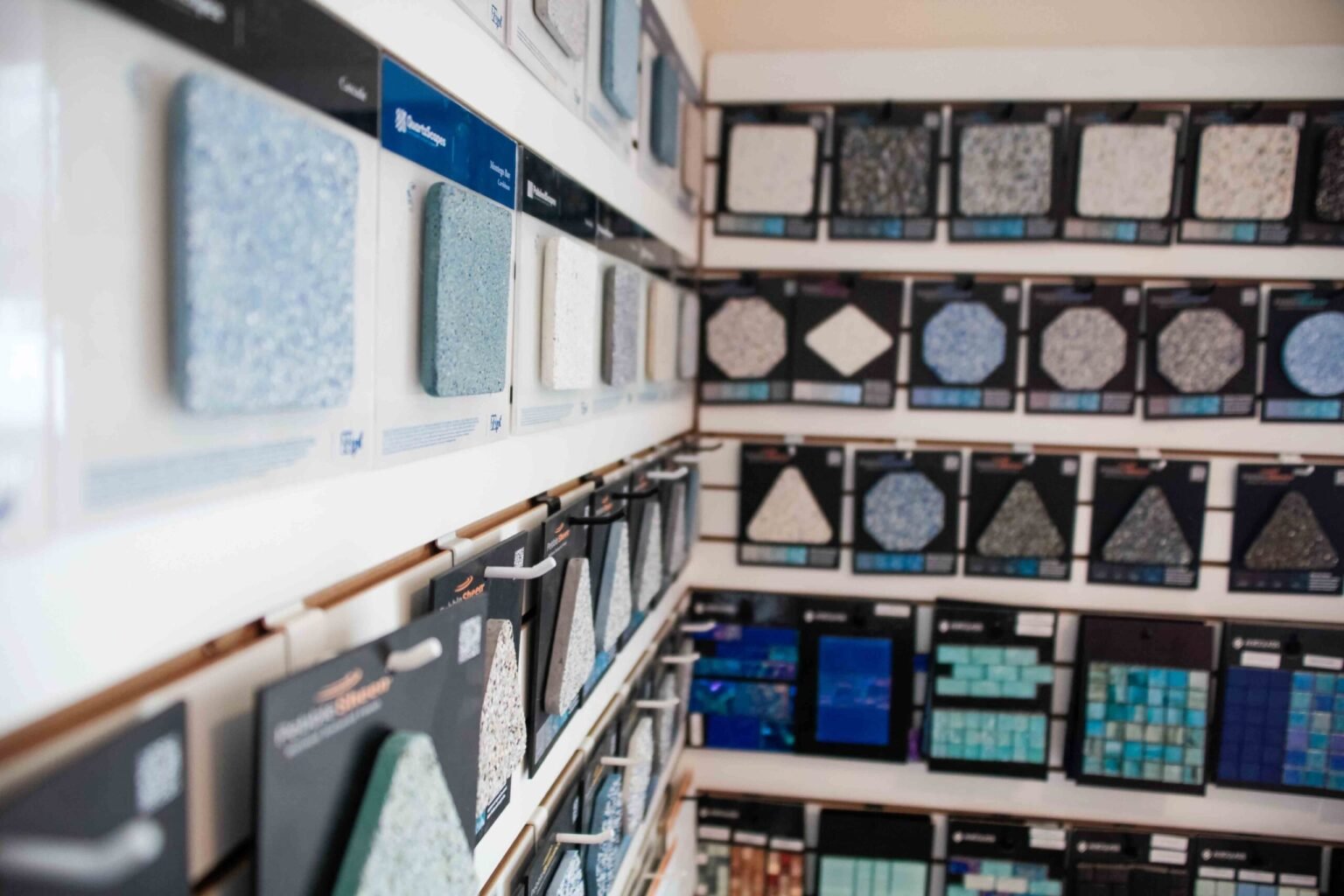 Indoor showroom display wall lined with pool finish and tile samples. Shelves hold plaster and aggregate finish samples in blues, grays, and whites alongside glass mosaic tile boards in teal, blue, and mixed tones, with brand labels including Quartzscapes 