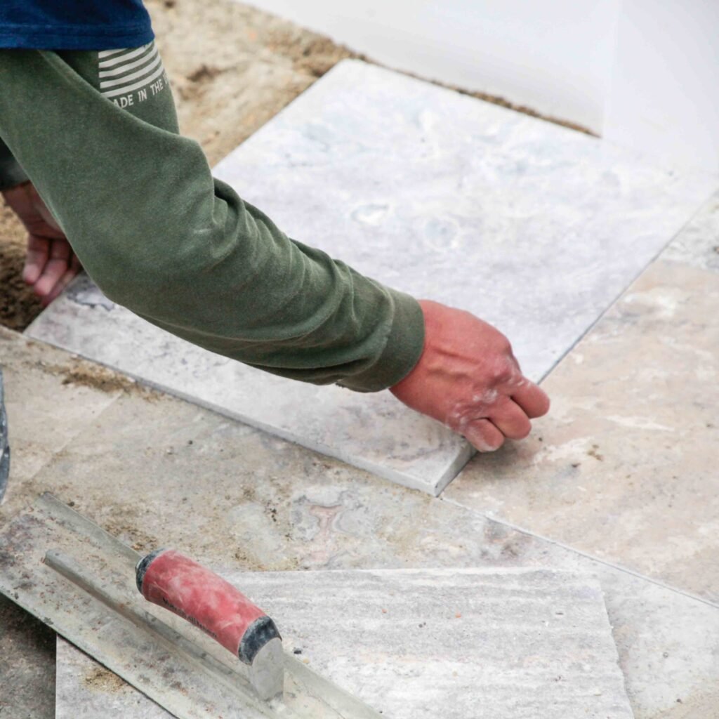 Construction photo showing a worker hand-setting large marble or travertine stone pavers on a sand base, with a rubber mallet trowel resting nearby on the partially completed surface.