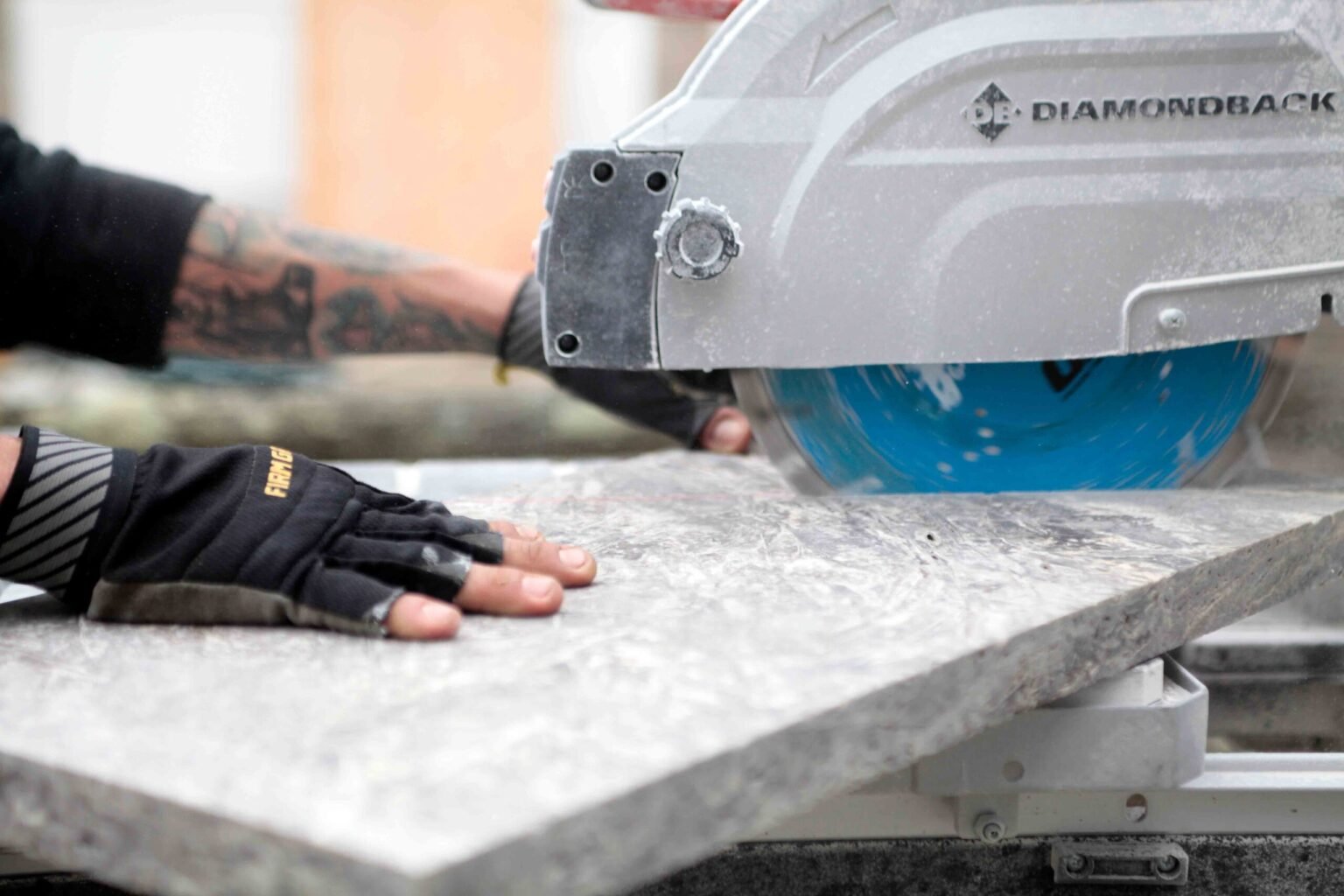 Construction process photo showing a worker's gloved hands guiding a large gray stone paver through a Diamondback diamond blade wet saw during a patio or pool deck installation.